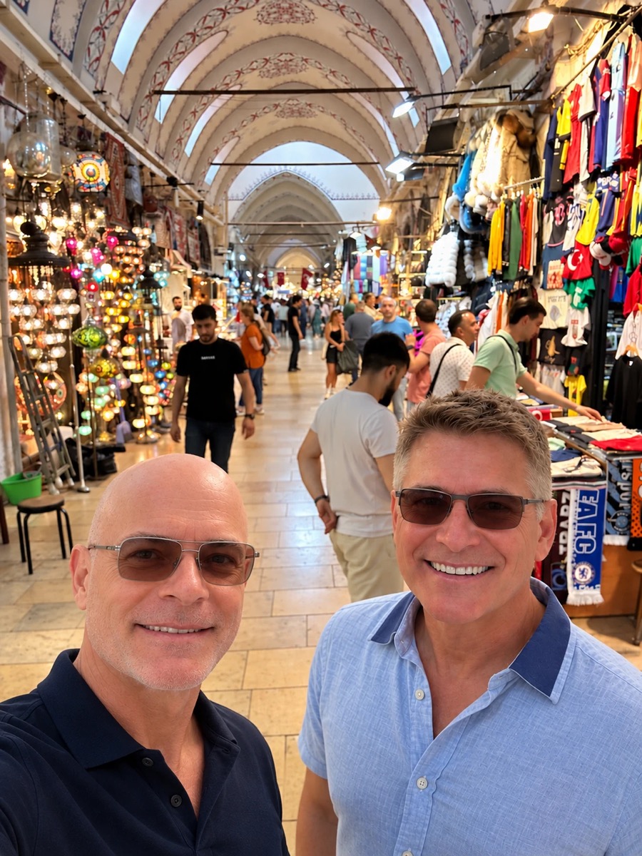 Two men smiling in a selfie inside the Grand Bazaar, under painted vaulted ceilings, with a wall of lit glass lamps on one side and hanging clothes on the other.